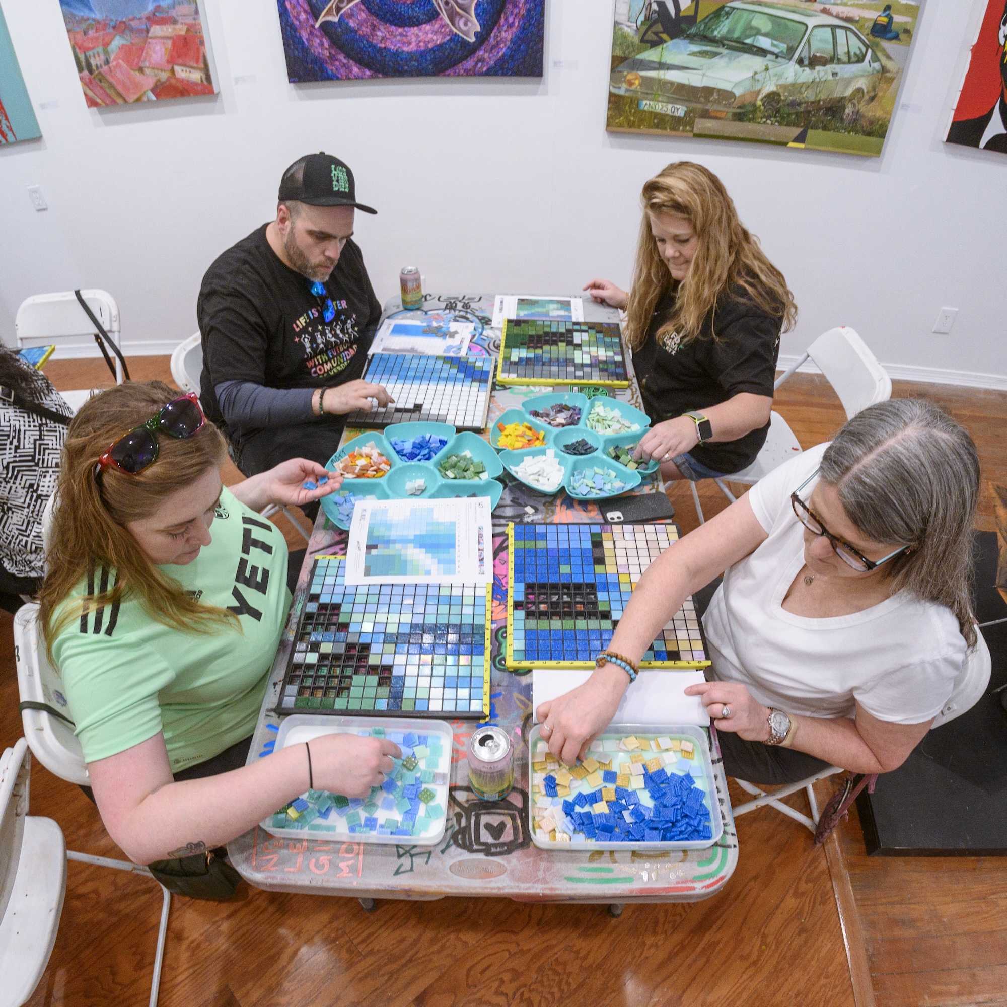 Four people sitting at a table, each with a tray of colorful tiles, filling a grid with 