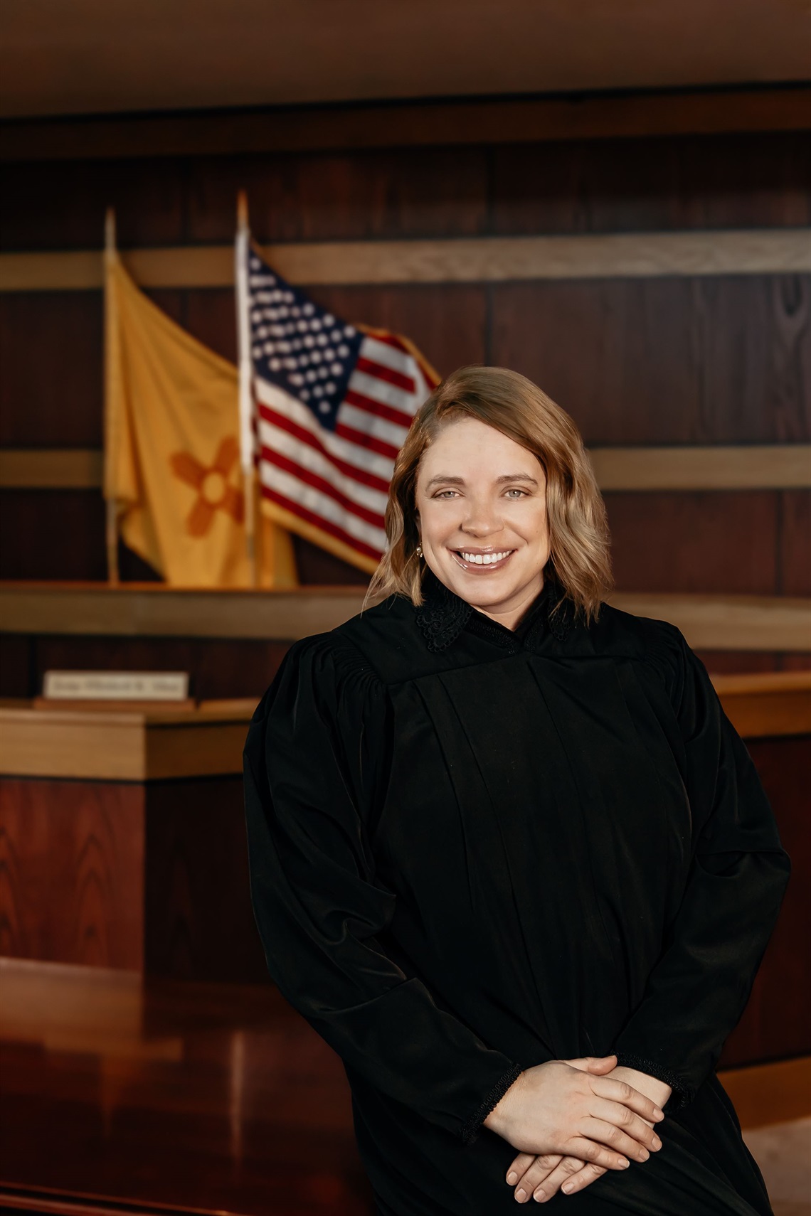A female judge in a black robe sits in front of an American flag and a New Mexico flag